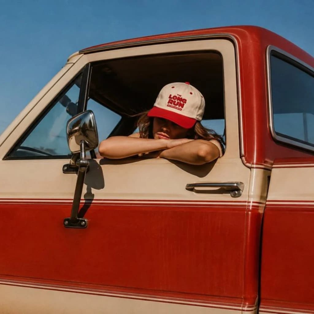 Person in a vintage red and cream truck wearing a red-brim The Long Run cap